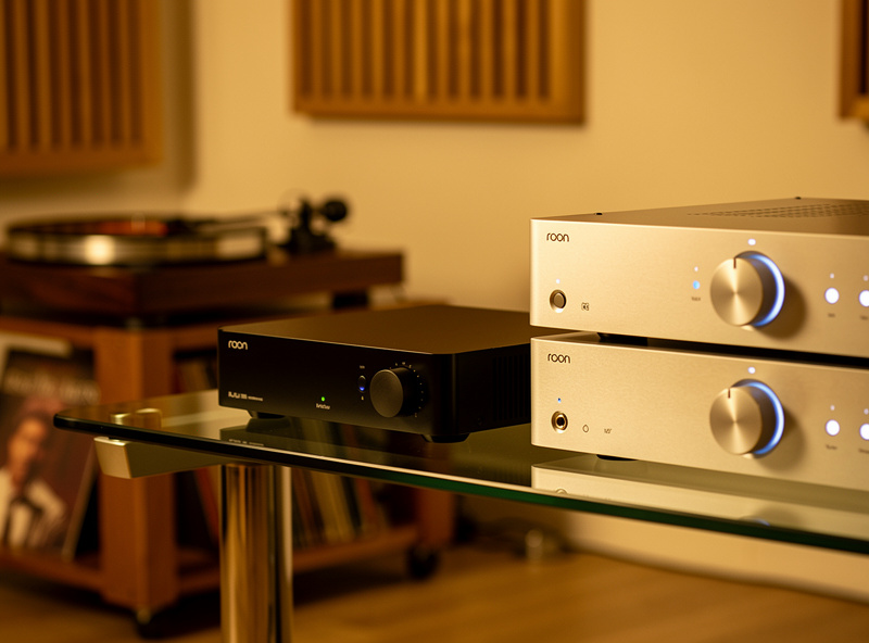 Roon Nucleus or similar music server appliance on glass shelf beside high quality DAC and pre-amplifier with turntable in background warm audiophile setup