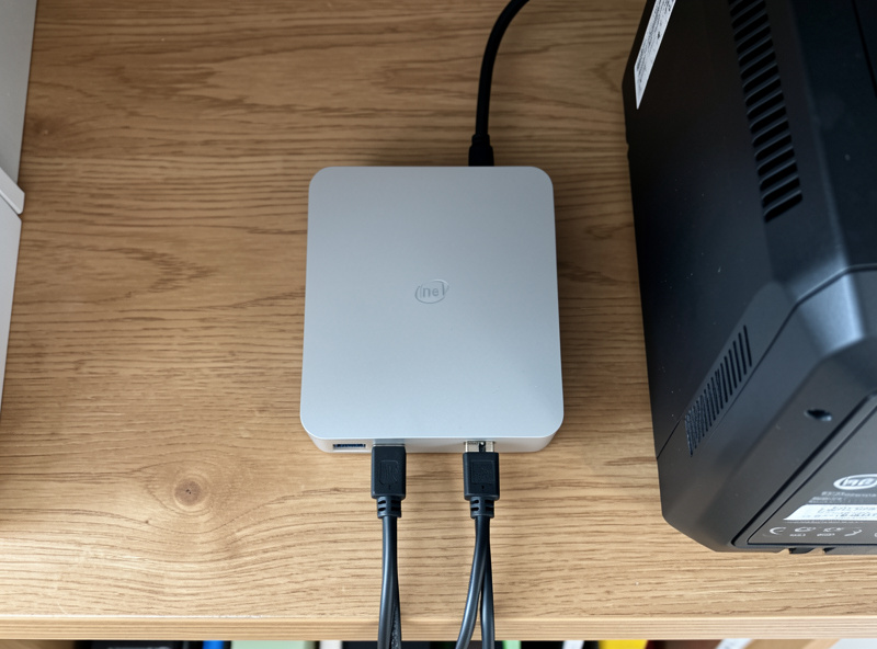 Fanless Intel N100 mini PC home server with two ethernet cables on a wooden shelf beside a UPS